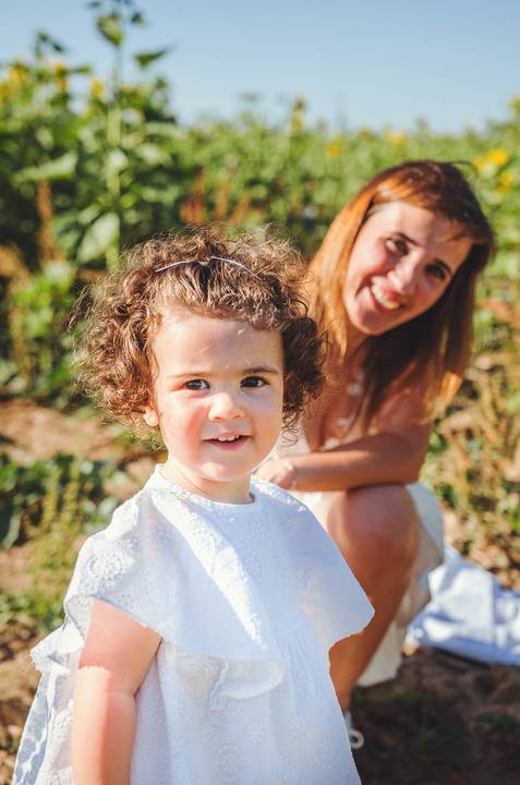 Close-up emocional de criança pequena sorrindo e olhando para câmera em sessão familiar. Menina em retrato individual com flores girassóis desfocadas ao fundo. Fotografia artística capturando beleza infantil em composição íntima.'