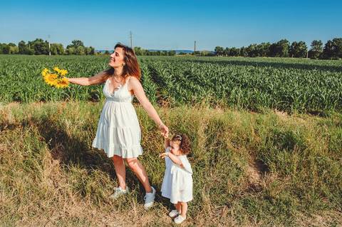 Mulher adulta com criança pequena em sessão familiar em ambiente rural aberto. Mãe e filha brincando juntas em campo com céu azul ao fundo. Fotografia profissional capturando vínculo familiar alegria em composição artística natural.'
