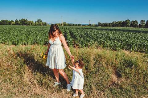 Mulher adulta e criança em retrato em sessão familiar ao ar livre em campo aberto. Mãe e filha brincando em ambiente rural natural com céu azul. Fotografia profissional capturando momentos familiares alegres em composição artística.'