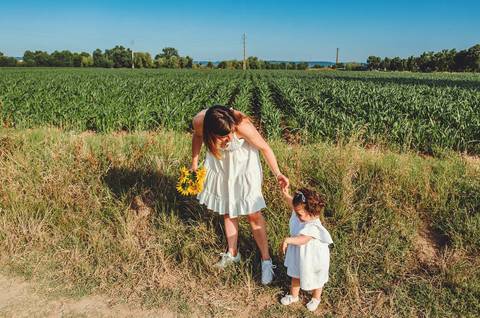 Mulher adulta com criança em retrato familiar em sessão ao ar livre em campo. Mãe agachada brincando com filha em ambiente rural natural. Fotografia profissional capturando momento emocional familiar alegre em composição artística.'