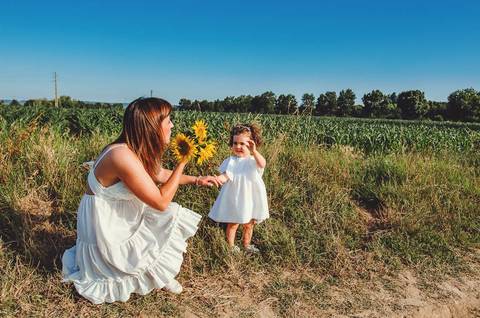 Mulher adulta agachada com criança pequena em sessão familiar ao ar livre. Mãe brincando com filha em ambiente rural natural de campo aberto. Fotografia profissional capturando vínculo familiar emocional em composição artística.'