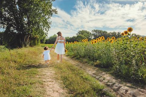 Mulher adulta em retrato familiar em sessão ao ar livre segurando criança pequena. Mãe em ambiente rural natural abraçando filha com céu azul ao fundo. Fotografia profissional capturando vínculo familiar emocional em composição artística natural.'