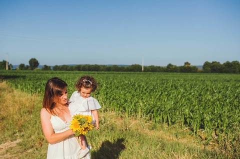 Mulher adulta com criança pequena em retrato familiar em sessão ao ar livre em campo. Mãe e filha brincando em ambiente rural natural com céu azul ao fundo. Fotografia profissional capturando vínculo familiar alegre em composição artística descontraída.'