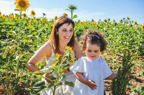 Sessão familiar em campo amplo de girassóis com paisagem natural ao fundo. Mulher e criança brincando juntas em ambiente rural idílico com céu azul. Fotografia profissional documentando momentos autênticos de família em estilo lifestyle outdoor.'