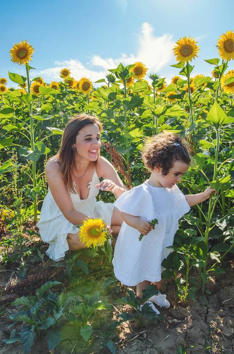 Mulher adulta e criança em retrato familiar durante sessão no campo de girassóis. Mãe segurando filha em ambiente rural com céu azul ao fundo. Fotografia profissional capturando momentos emocionales de vínculo familiar em composição artística.'