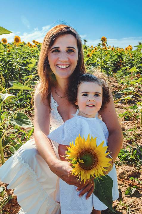 Retrato emocional de mãe e filha durante sessão familiar no campo de girassóis. Mulher adulta com criança pequena em momento afetuoso e natural. Fotografia profissional capturando vínculo familiar em ambiente rural com iluminação solar natural.'