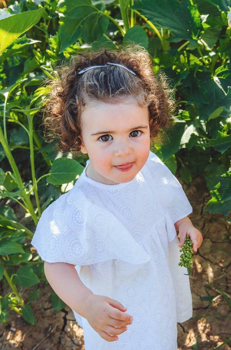 Menina em close-up emotivo rodeada de vegetação verde e vibrante durante sessão fotográfica ao ar livre. Retrato natural de criança em campo de girassóis, capturando a inocência e beleza infantil. Fotografia profissional com excelente iluminação natural.'