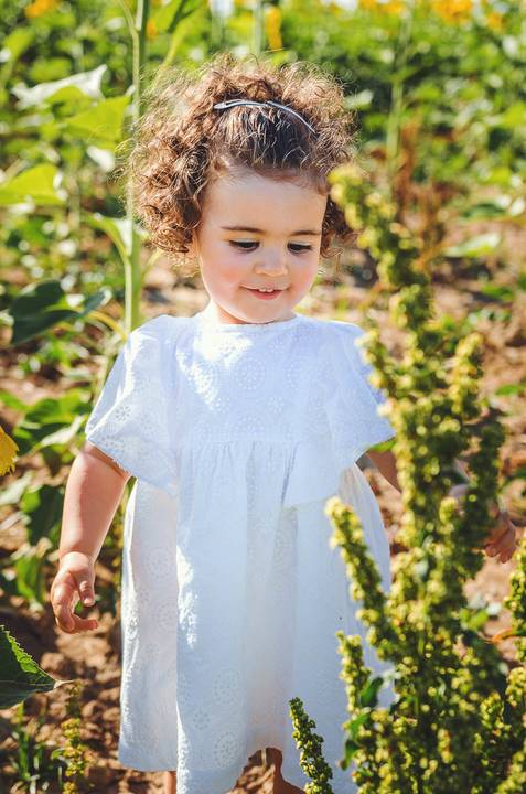 Menina pequena em retrato individual em ambientes naturais durante sessão fotográfica profissional ao ar livre. Criança brincando em campo de girassóis com expressão natural alegre. Fotografia artística capturando beleza infantil em composição cuidadosa e'