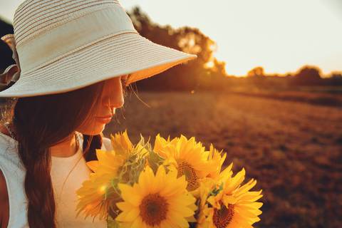 Mulher com chapéu segurando girassol iluminada pelo sol poente'