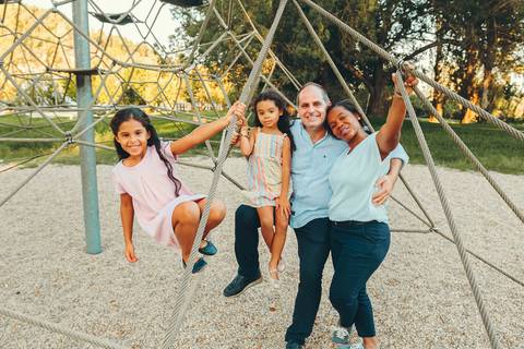 Família brincando e correndo em parque infantil durante sessão fotográfica, criança subindo em estrutura de lazer com pais acompanhando, momento espontâneo de diversão ao ar livre'