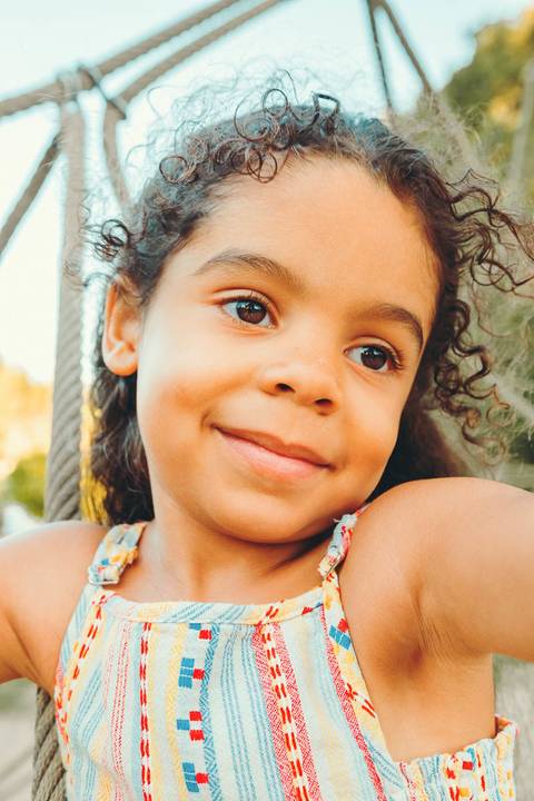 Menina pequena em close com sorriso durante sessão fotográfica de família, retrato infantil com luz natural ao entardecer, fotografia de criança alegre em estúdio outdoor'