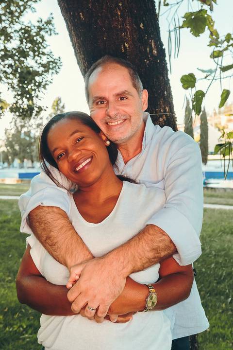 Casal de pais abraçados e sorrindo durante sessão fotográfica de família em parque ao entardecer, retrato de casal com abraço carinhoso à beira da água com luz natural golden hour'