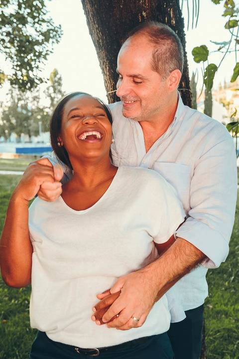 Casal sorrindo juntos durante sessão fotográfica de família em parque ao entardecer, retrato de cônjuges com abraço caloroso na beira do rio com luz dourada ao pôr do sol'
