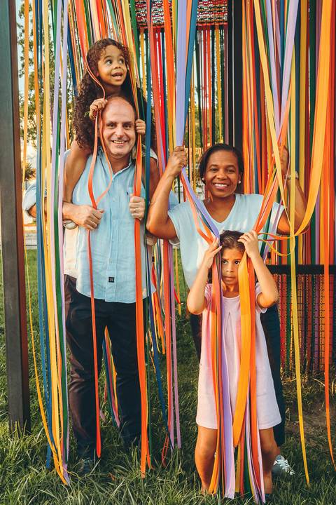 Família posando em frente a estrutura de tecido colorido em parque, sessão fotográfica de família ao ar livre com filha em primeiro plano, retrato familiar descontraído com luz natural'