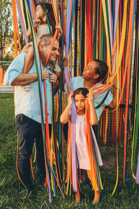 Família posando em frente a tecido colorido com filha abraçada à mãe, sessão fotográfica de família em estrutura de parque ao ar livre, retrato familiar afetuoso com luz natural'