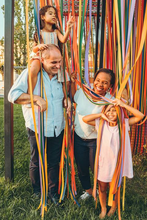Família posando junto a árvore em caminho de parque ao entardecer, sessão fotográfica ao ar livre com luz golden hour, retrato familiar ao caminhar em ambiente natural verde'