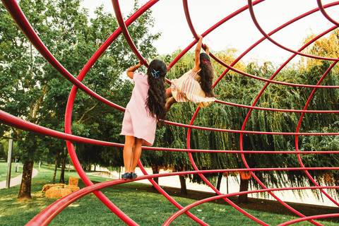 Menina pendurada em estrutura vermelha de parque ao pôr do sol durante sessão fotográfica, retrato infantil brincando em equipamento de lazer ao ar livre com luz golden hour natural'