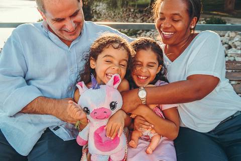 Pai brincando com filhas pequenas, momento espontâneo durante sessão fotográfica de família ao ar livre, interação genuína de pais e filhos em ambiente natural e aconchegante'