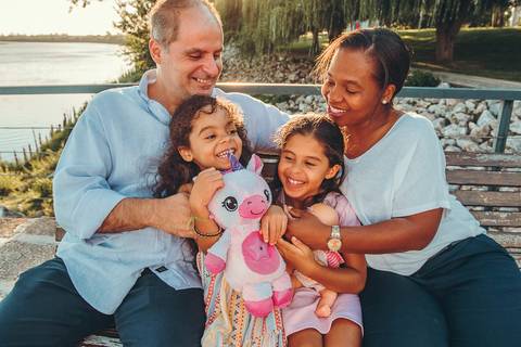 Pai abraçando filhas sentadas em pedras próximo à água, sessão fotográfica de família com vista de paisagem natural, momento íntimo e afetuoso de pais e filhos ao ar livre'