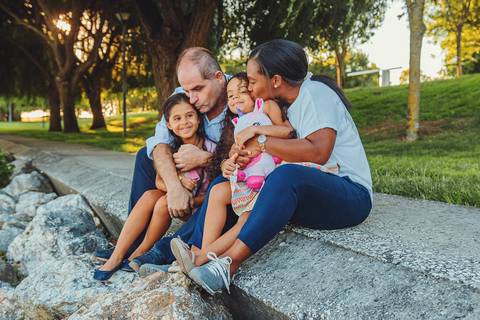 Família sentada em grama verde durante sessão fotográfica ao ar livre, momento descontraído de pais com filhas em parque natural com luz golden hour ao pôr do sol'