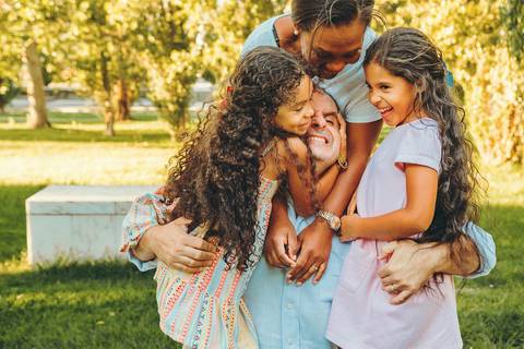 Mãe sorrindo com filhas ao seu redor durante sessão fotográfica em ambiente de parque com árvores, fotografia de família com luz natural ao entardecer em Vila Nova da Barquinha'