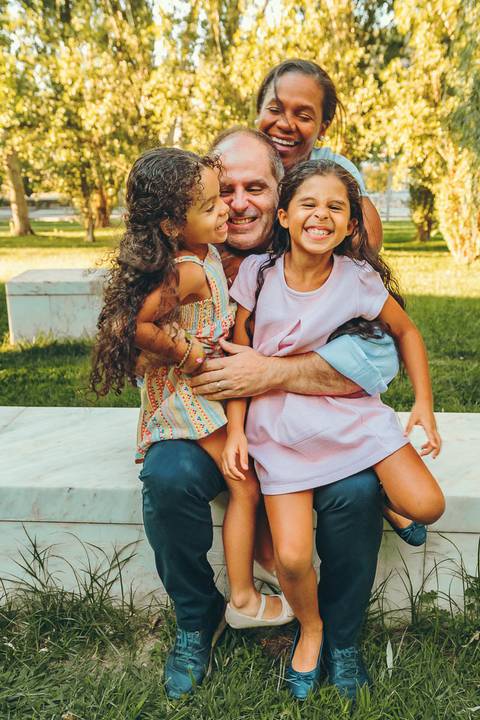 Mãe com duas filhas abraçadas em ambiente ao ar livre com grama verde, sessão fotográfica familiar calorosa com luz dourada ao pôr do sol, retrato lifestyle de mãe e filhos'