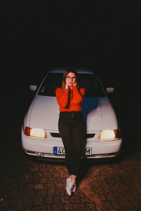 Myriam Correia de look laranja com o Seat Ibiza iluminado ao fundo durante retrato noturno na Quinta da Cardiga, Golegã. Pose confiante com atmosfera cinematográfica. Duo Capuano Fotografia.'