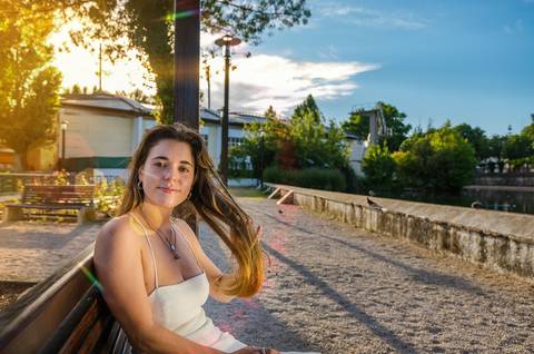 Susana sentada ao pôr do sol junto ao lago com luz quente e dourada. Expressão descontraída e vestido branco. Fotografia feminina individual em Torres Novas ao entardecer por Duo Capuano.'
