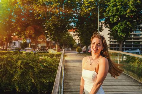 Susana apoiada em corrimão metálico ao pôr do sol em Torres Novas. Vestido branco fluido e expressão serena. Fotografia feminina individual ao entardecer no parque urbano por Duo Capuano.'