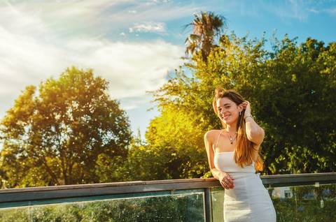 Susana apoiada em varanda metálica junto ao lago ao entardecer, vestido branco e sorriso aberto. Cenário verde e palmeiras ao fundo. Sessão fotográfica individual em Torres Novas por Duo Capuano.'
