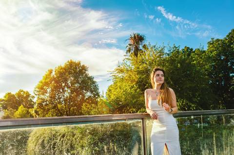 Susana em vestido branco com pose elegante junto ao lago ao entardecer. Céu nublado com tons dourados e vegetação exuberante ao redor. Sessão fotográfica feminina individual por Duo Capuano.'