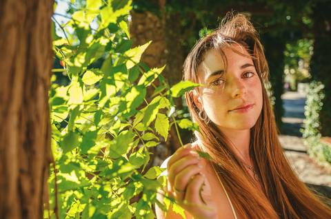 Retrato feminino a contraluz com folhagem verde ao fundo. Susana com expressão suave e olhar introspetivo. Fotografia individual ao ar livre no Jardim das Rosas, Torres Novas, Duo Capuano.'