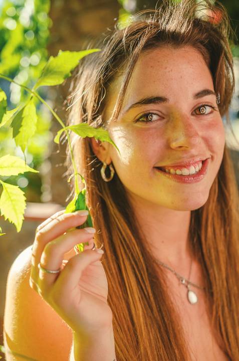Retrato feminino em close com sorriso espontâneo e luz dourada. Susana segura folhagem verde enquanto posa descontraidamente. Sessão individual no Jardim das Rosas, Torres Novas, Duo Capuano.'