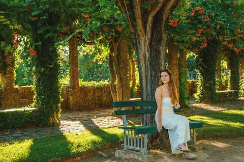 Susana sentada em banco de jardim sob sombra de árvores floridas no Jardim das Rosas, Torres Novas. Pose relaxada em vestido branco. Fotografia feminina individual por Duo Capuano Fotografia.'