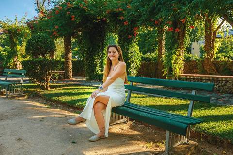 Susana sentada em banco de madeira no jardim com fundo verde desfocado. Pose descontraída em vestido branco. Sessão fotográfica feminina individual no Jardim das Rosas, Torres Novas, Duo Capuano.'