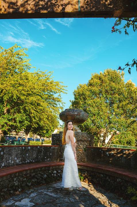 Fotografia feminina em jardim público de Torres Novas com Susana em vestido branco e céu azul ao fundo. Pose elegante junto à pedra histórica. Sessão individual ao ar livre por Duo Capuano.'