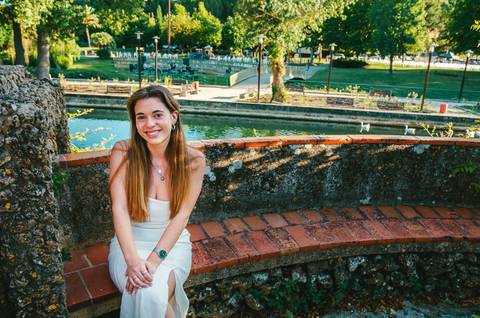 Susana sentada junto a lago com reflexo de céu azul e vegetação exuberante. Vestido branco e sorriso sereno. Fotografia feminina individual em Torres Novas por Duo Capuano Fotografia.'