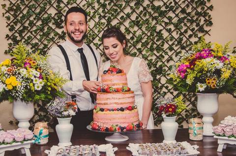 Casal Camila e Rafael cortando bolo de casamento em momento festivo durante festa em São Paulo com mãos juntas'