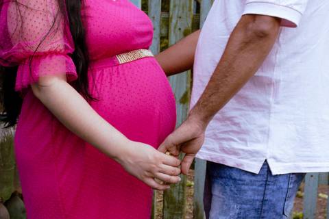 Barriga de Gestante com vestido cor de rosa. Casal segurando a mão durante o ensaio'