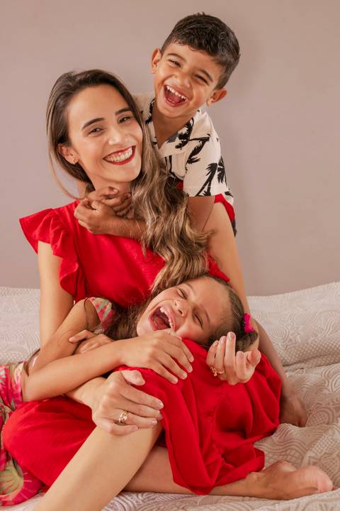 Mãe sentada na cama enquanto o filho a abraça pelo pescoço e a filha está deitada no colo'