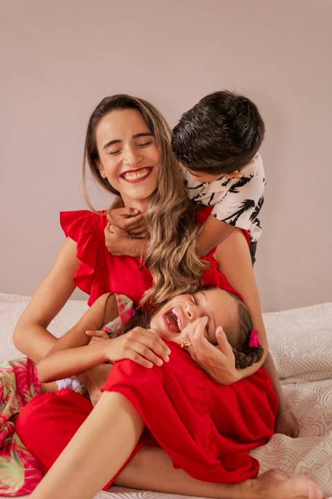 Mãe sentada na cama enquanto o filho a abraça pelo pescoço e a filha está deitada no colo'