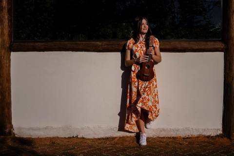 Garota com vestido de estampa laranja apoiada numa parede branca com um dos pés segurando um UKULELE enquanto posa para foto de Mary Santtiago, fotógrafa de Suzano SP'