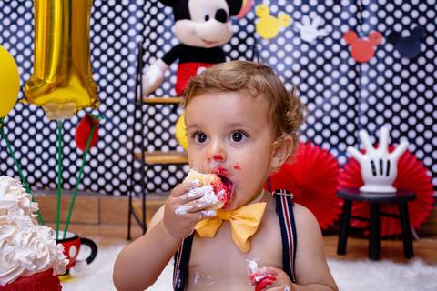 O bebê come um pedaço de bolo com as mãozinhas cheia de coberto do bolo do Mickey Mouse, no seu Ensaio Smash the Cake feito em Suzano'