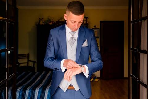 Um noivo com um fato azul ajusta seu relógio enquanto está em uma sala com piso de madeira. Ele veste um colete cinza claro e gravata, com um lenço de bolso. O quarto tem uma cama com um cobertor listrado e uma porta ao fundo.'