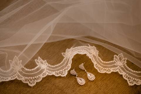 Um delicado véu de renda branca está estendido sobre uma superfície castanha, com um par de elegantes brincos de cristal em forma de lágrima colocados no topo. A renda apresenta padrões florais ao longo das bordas.'