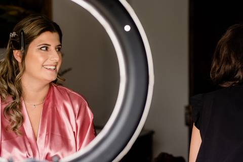 Uma noiva com um robe rosa, com cabelo estilizado preso de um lado, está sorrindo enquanto está sentada perto de um anel de luz brilhante. O cenário é uma área de preparação  para um casamento.'