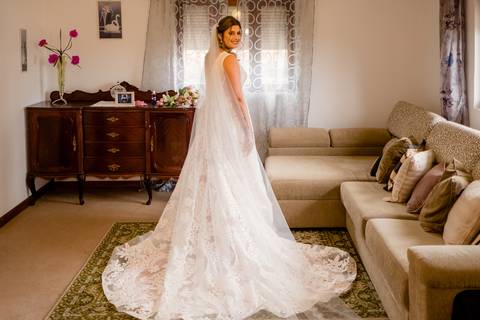 Uma noiva com um vestido de noiva de renda branca e véu longo está em uma sala de estar com um sofá bege e aparador de madeira. Ela está sorrindo e olhando para trás por cima do ombro. A sala é decorada com cortinas florais e fotos emolduradas.'