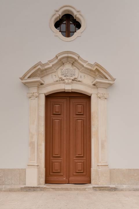   na Igreja de Portunhos Uma grande porta de madeira ornamentada com painéis intrincados em uma moldura de pedra clara contra uma parede branca. Acima da porta há uma pequena janela redonda e um arco de pedra decorativo com a data 1785 gravada.'