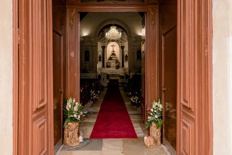 Portas de madeira abertas levam a uma igreja com um corredor de tapete vermelho. O interior é iluminado por um lustre, com arranjos florais perto da entrada. Um altar com uma cruz é visível ao fundo.'