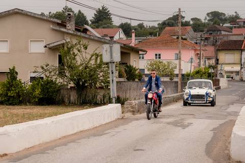 Uno Noivo guia a sua moto vintage por uma rua residencial com casas suburbanas ao fundo. Um carro clássico segue atrás. A cena tem um céu nublado.'
